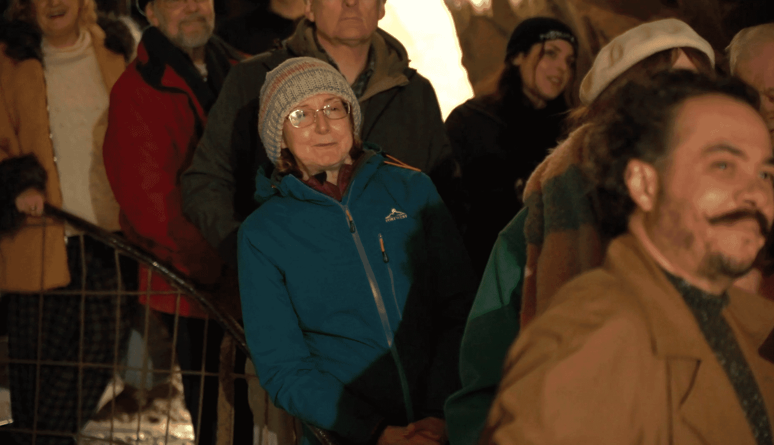 A woman in a blue fleece jacket and a light blue hat sits in a crowd and leans in to watch what is on stage