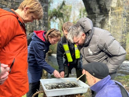 a group of people in outdoor coats gather around a water sampling tray to look at what is inside