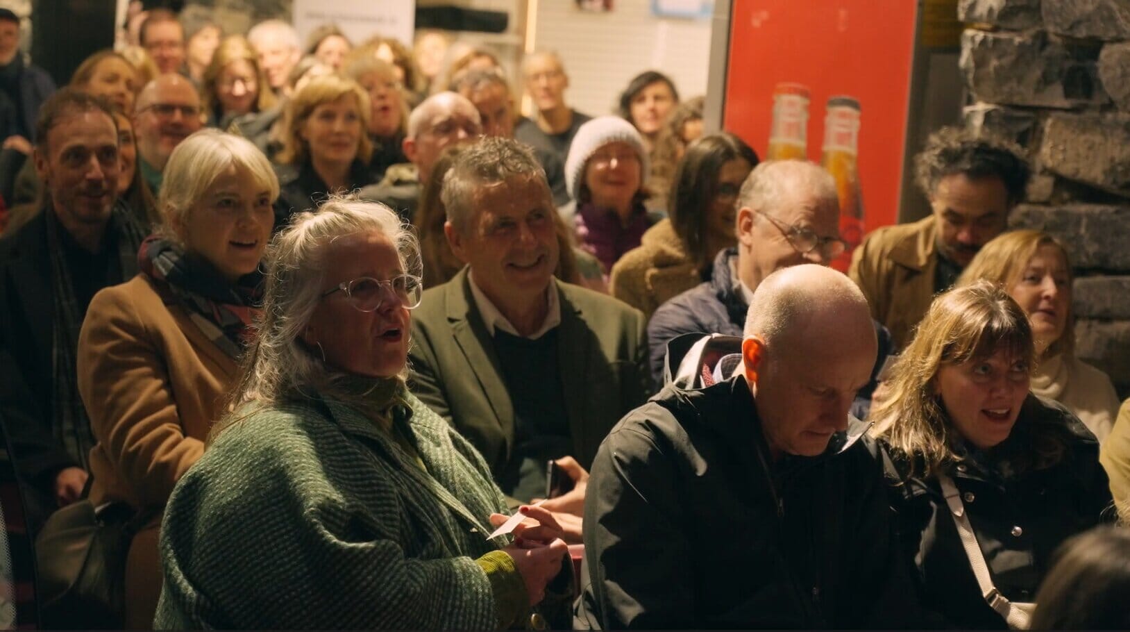 A group of people sit and stare at a stage ahead behind the camera