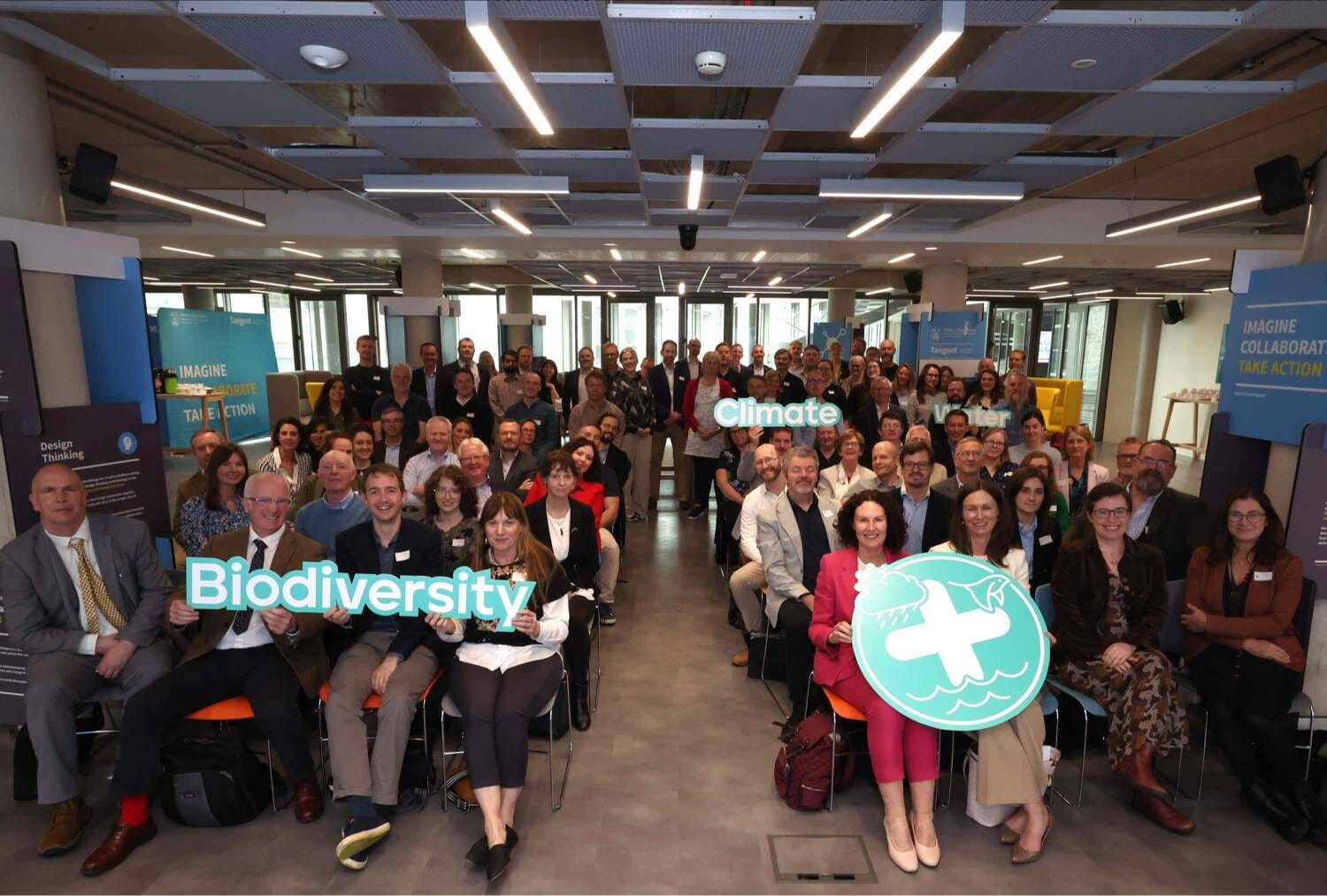 A group of people seated in a room holding signs that say climate, biodiversity and water.