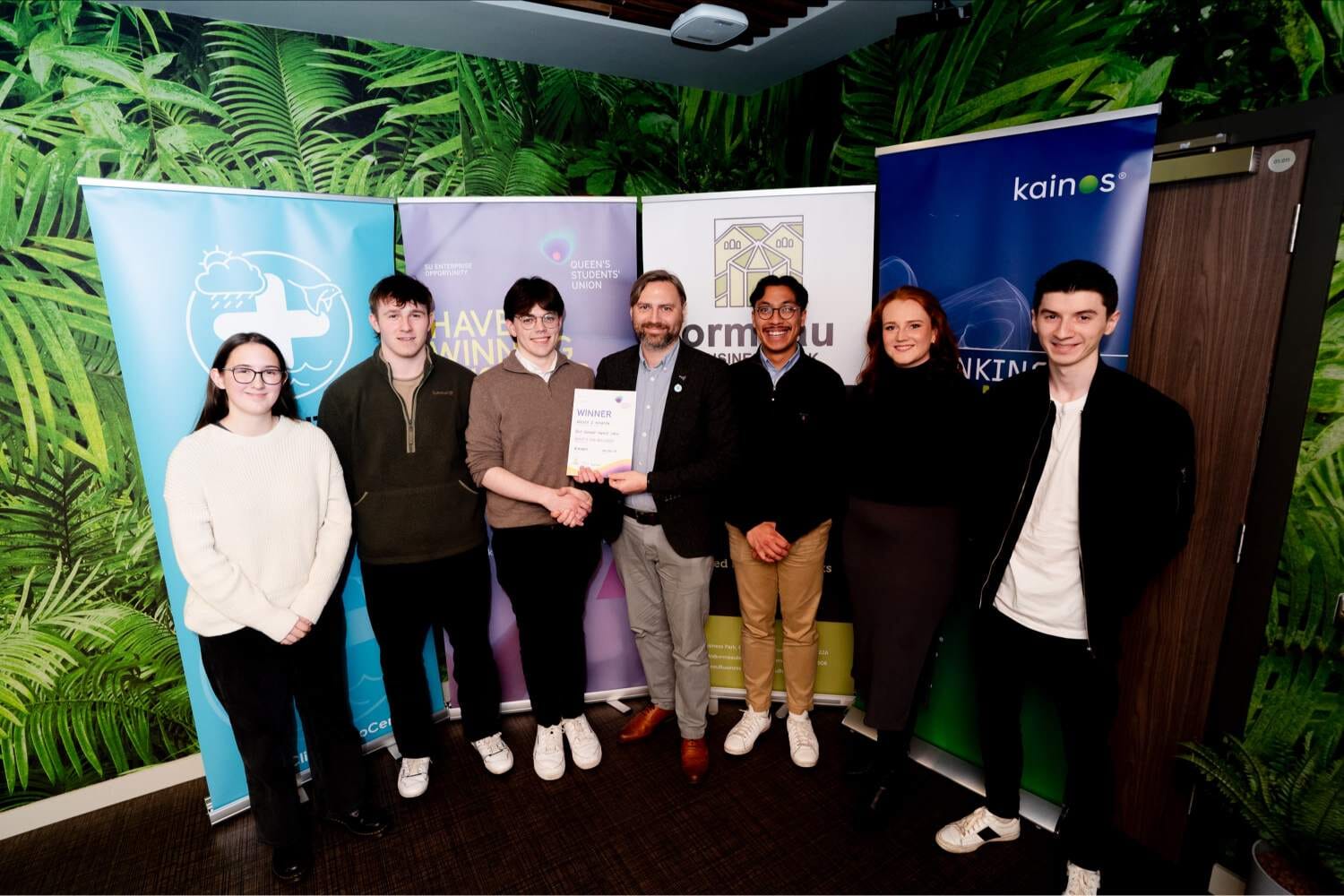 7 people stand in front of colourful pull up banners and get presented with an award